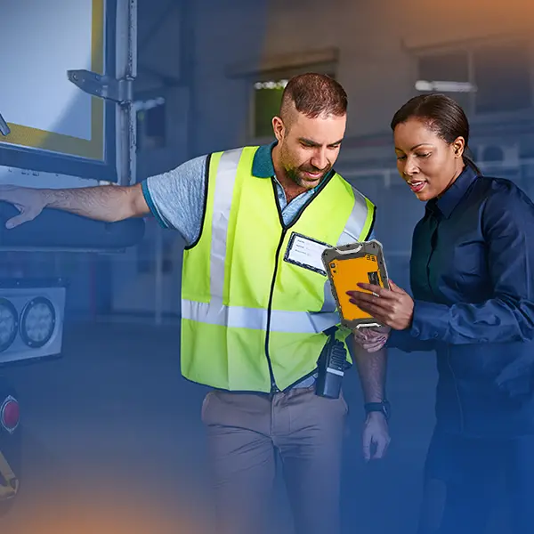 Two workers in a warehouse setting review information together on a tablet near a loading dock door.
